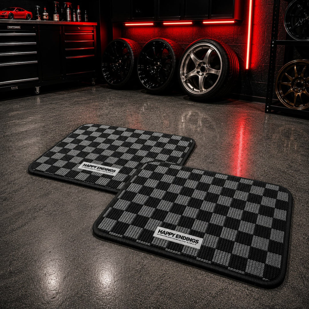 Two checkered car mats labeled HAPPY ENDINGS on a garage floor, with car whee...