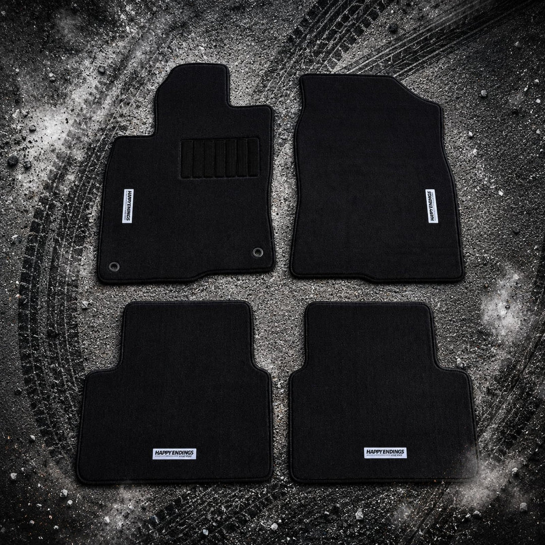 Four black car floor mats on a dark, textured surface with visible tire track...
