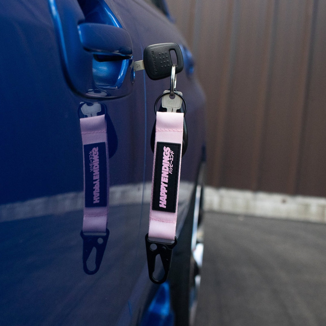 Car key with a pink Happy Heads keychain hanging from a blue car door.