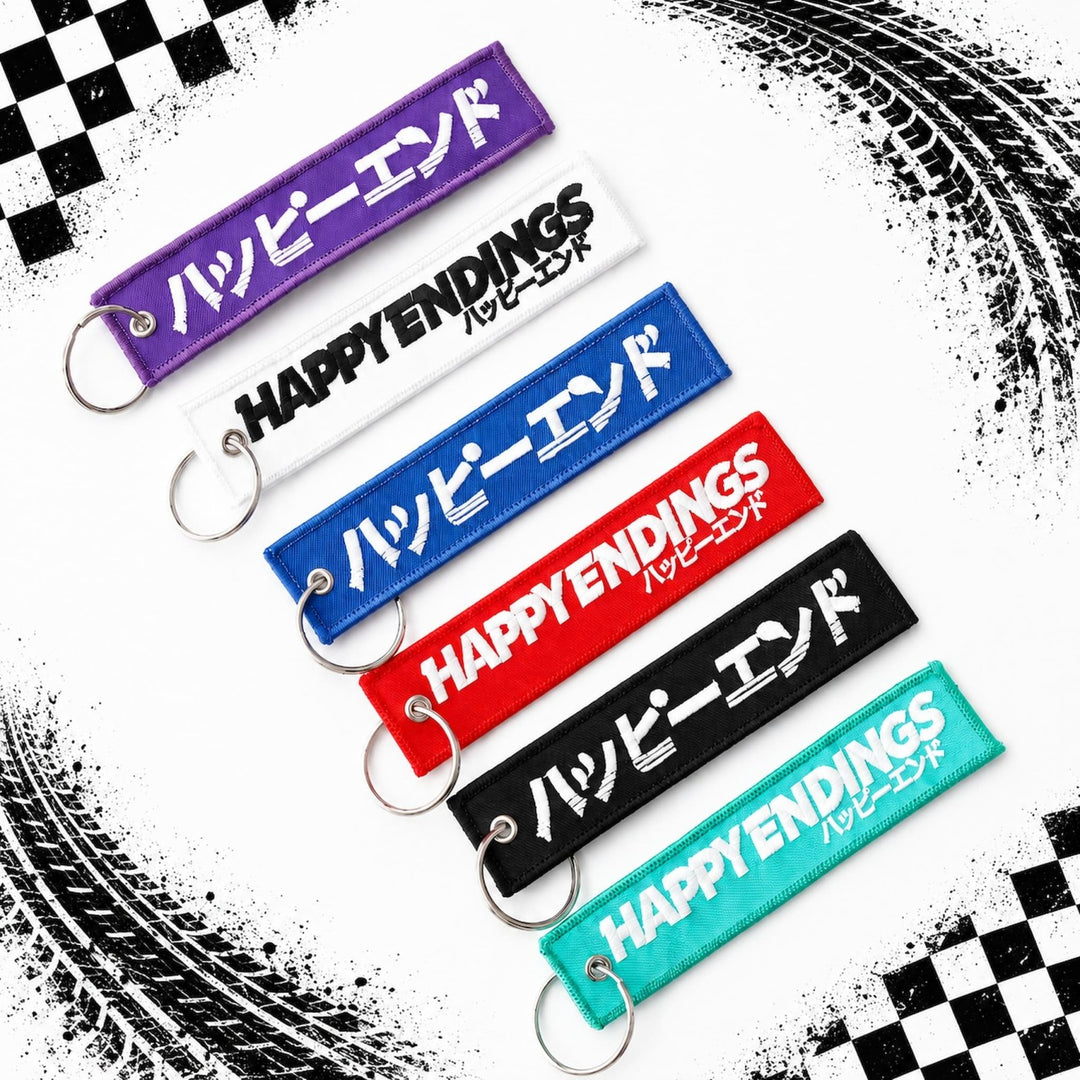 Six HAPPY ENDINGS keychains in different colors, shown on a tire-marked backg...