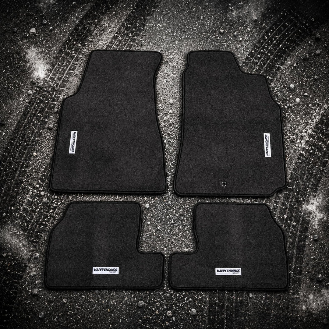Four black car floor mats arranged on gravel with visible tire tracks and Hap...
