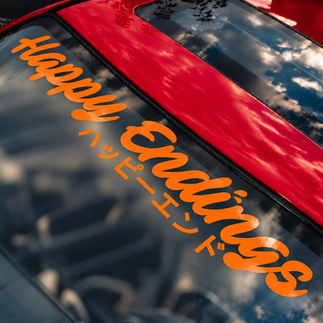 Orange decal reading Happy Endings in English and Japanese on a red car windshield.