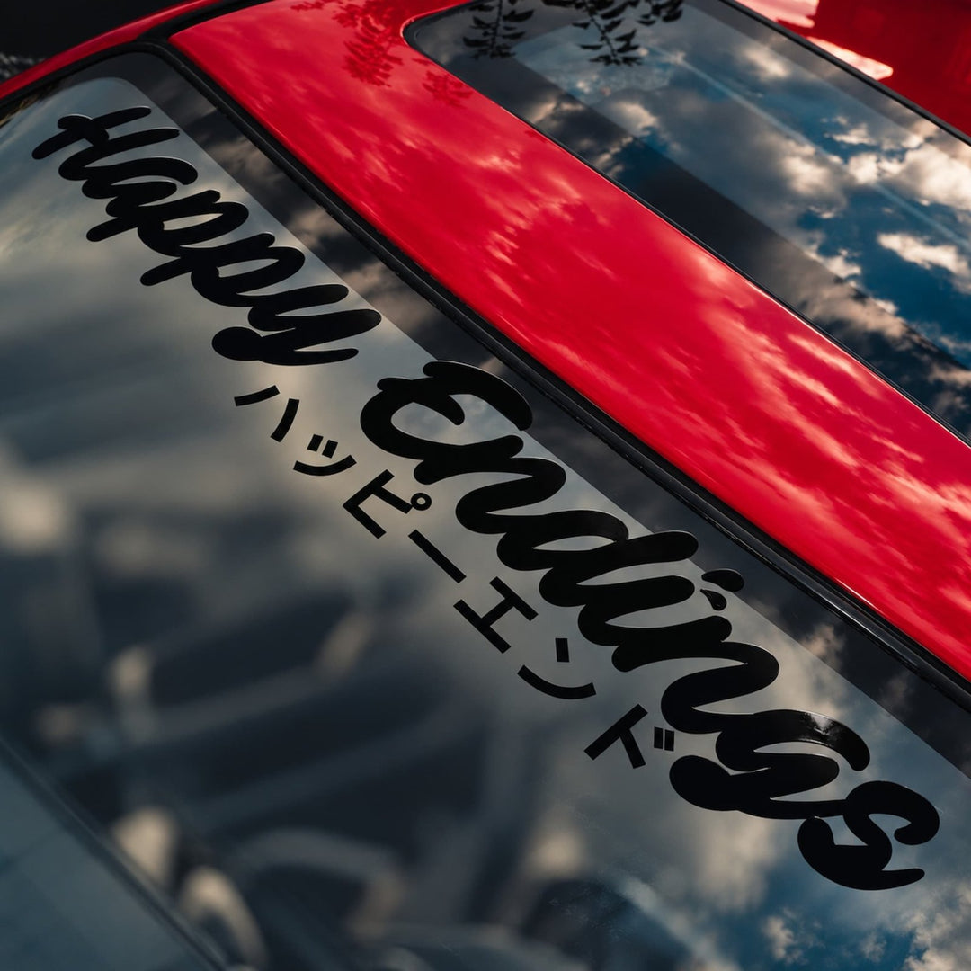 A red car windshield with a black decal that reads Happy Endings in English and Japanese.
