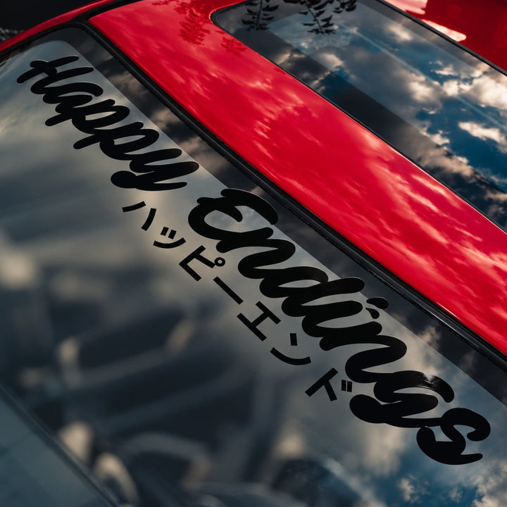 A red car windshield with a black decal that reads Happy Endings in English and Japanese.
