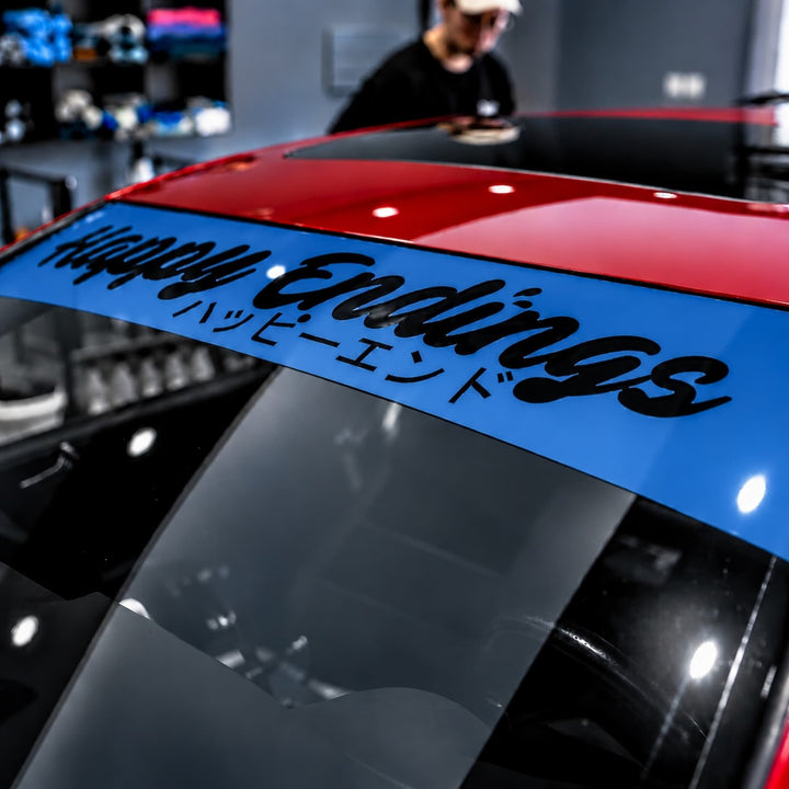 Blue windshield banner on a red car with the text Happy Endings in English and Japanese.