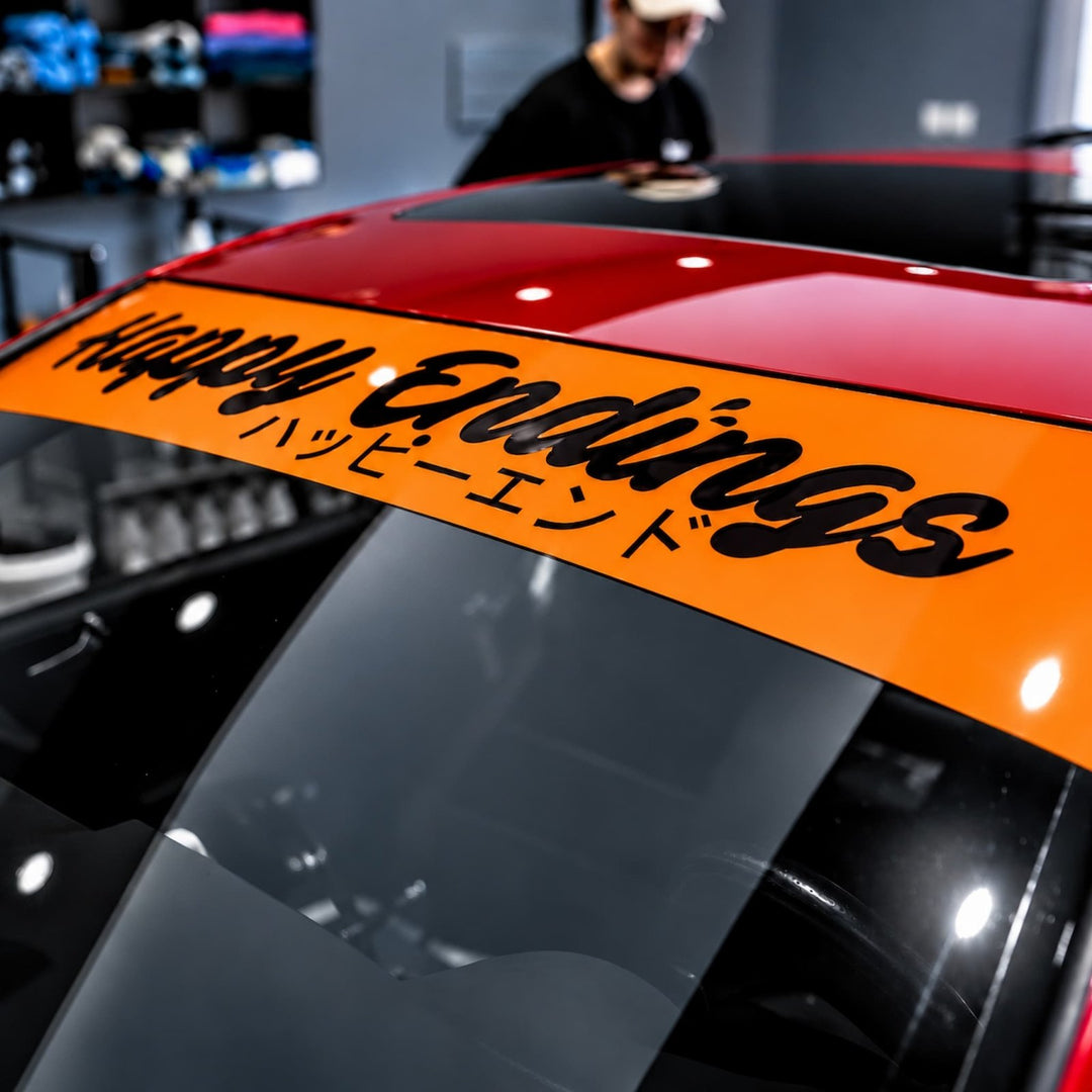 Orange windshield banner on a red car that reads Happy Endings in English and Japanese.