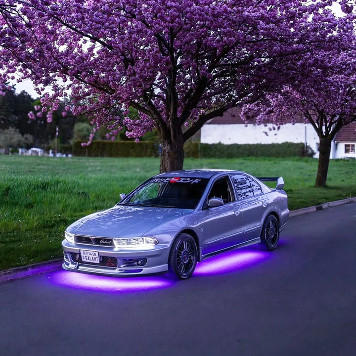 Silver car with purple underglow parked under blooming cherry blossom trees along a quiet street.