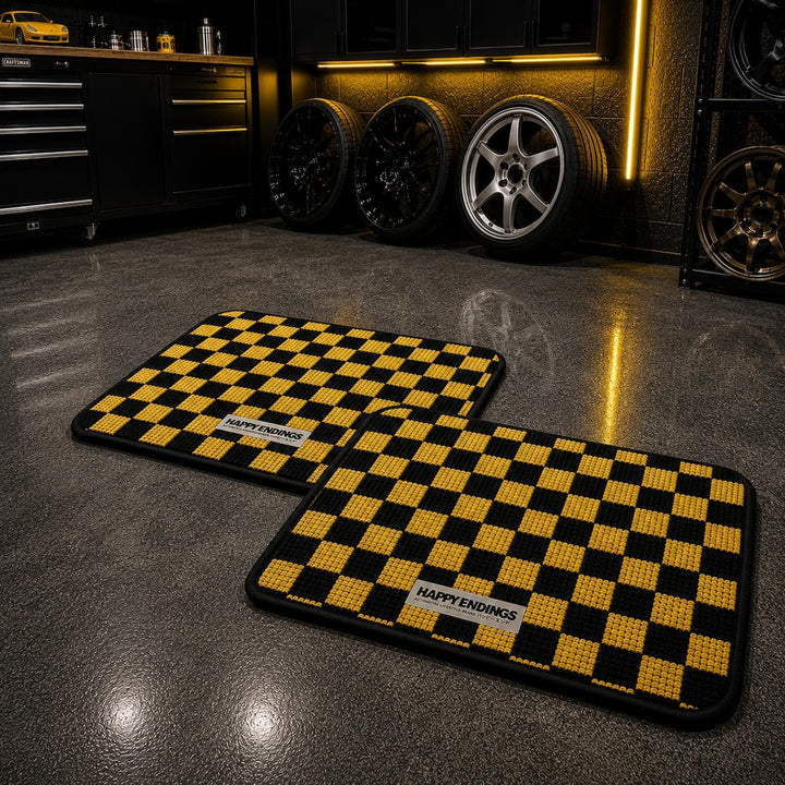 Two yellow and black checkered floor mats labeled HAPPY ENDINGS in a sleek garage with alloy wheels on the wall.